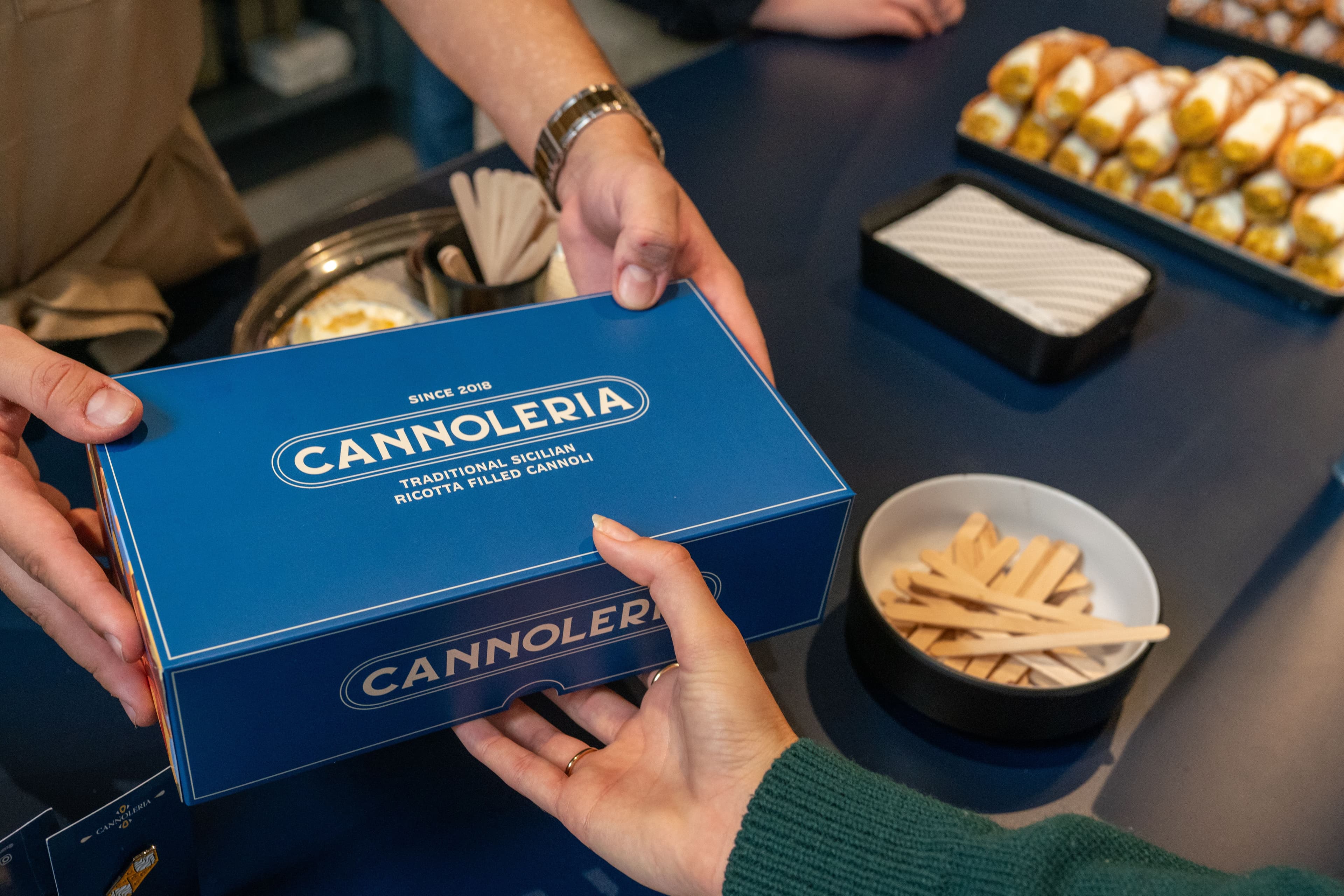 Hands passing blue Cannoleria box with cannoli tray behind