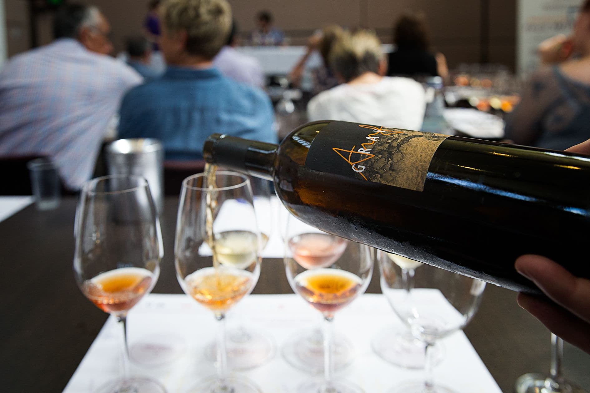Wine bottle pouring into glasses at tasting event with attendees in background