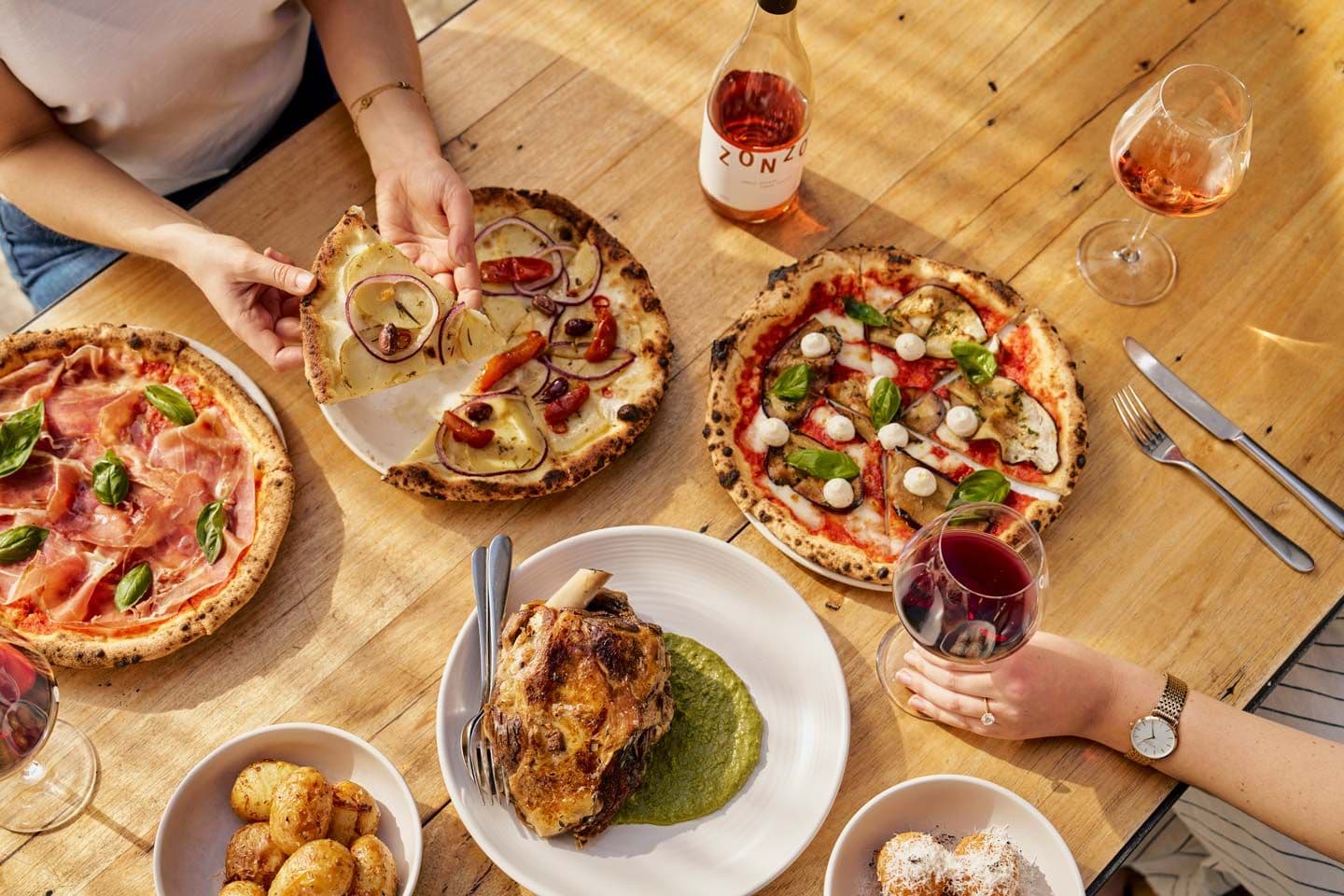 Wood-fired pizzas, wine glasses and roast dish on wooden table