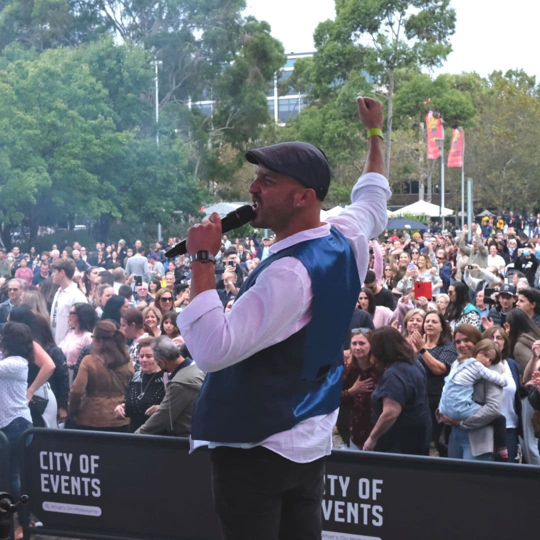 Male performer singing into microphone on stage, fist raised, large crowd behind barrier