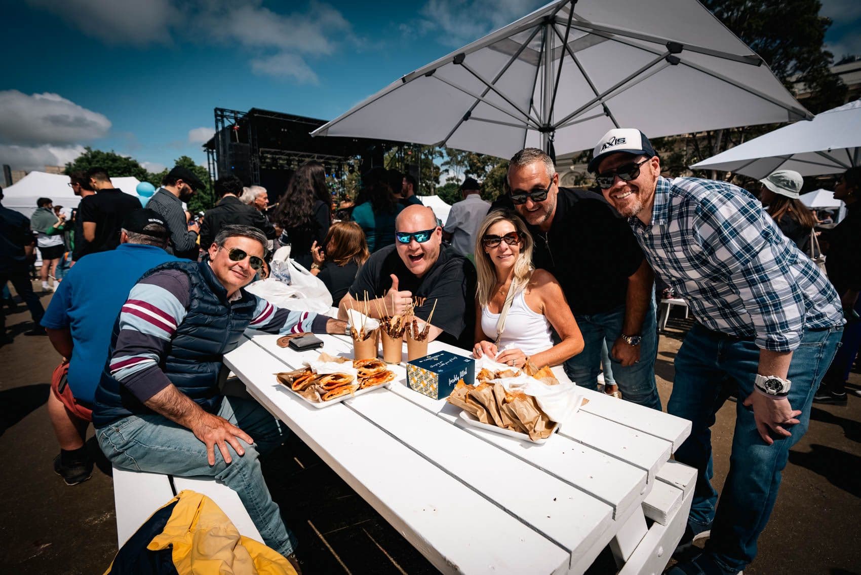 Group of smiling guests enjoying food and drinks at a shaded outdoor festival table