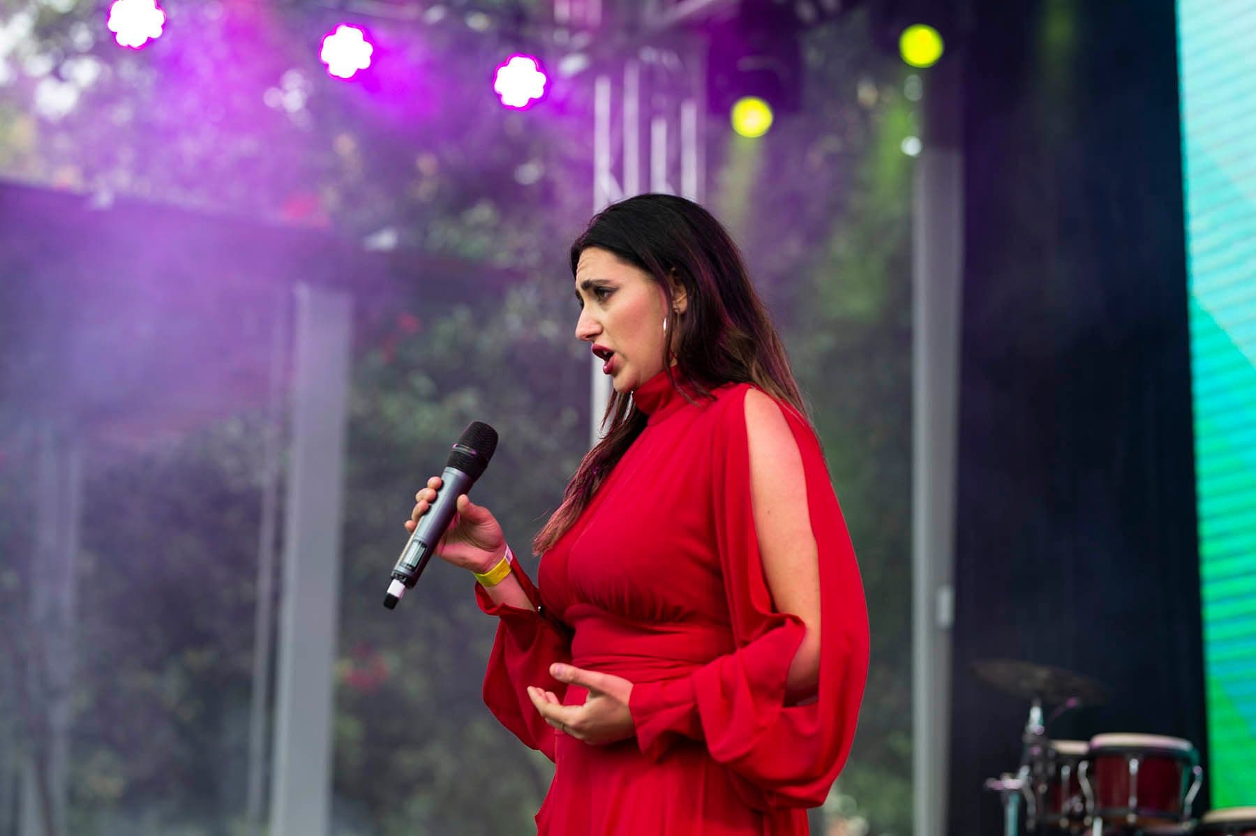 Female performer in red dress singing with microphone on stage with purple and teal lighting