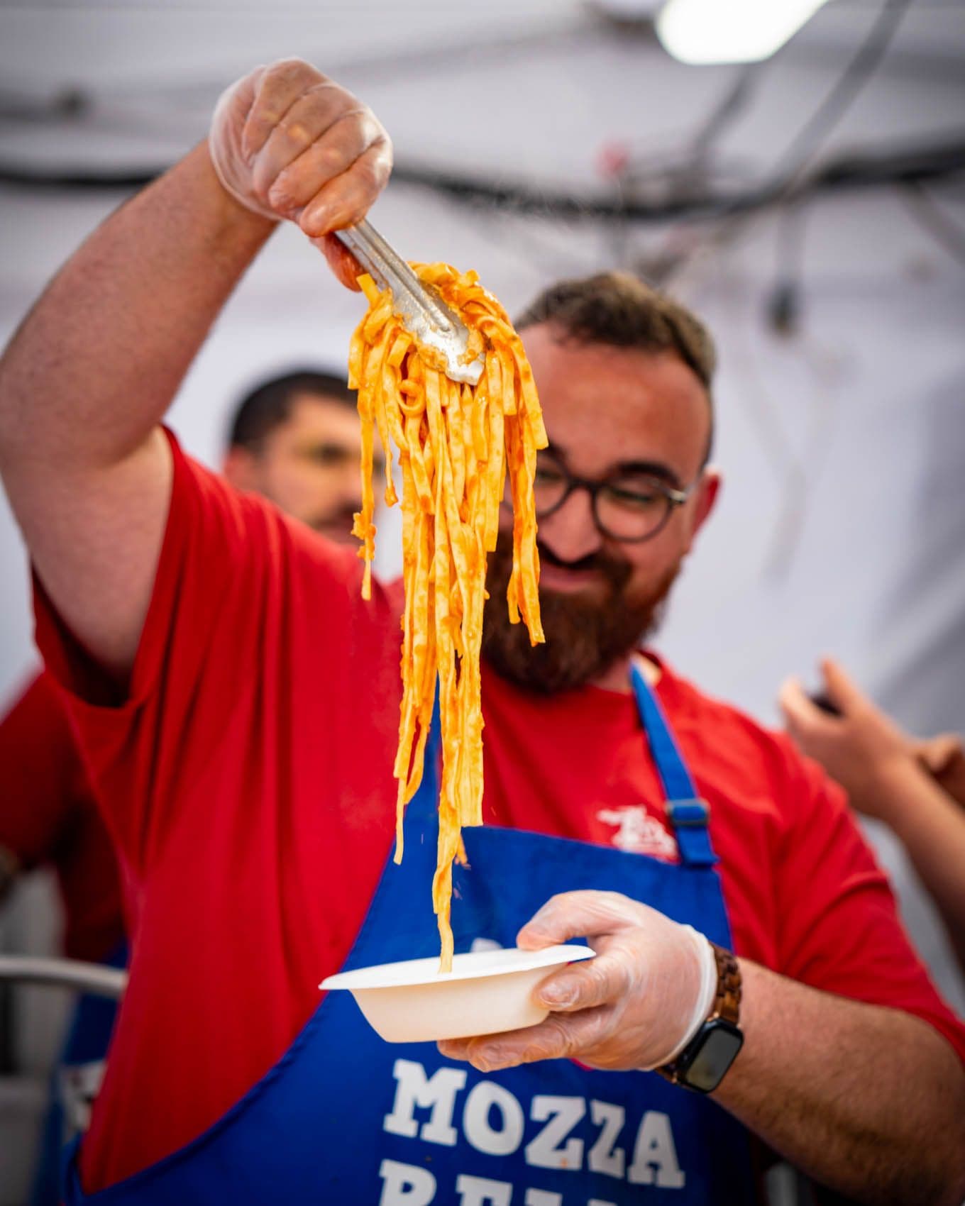 Chef lifting fresh pasta on fork, wearing red shirt and blue Mozza apron