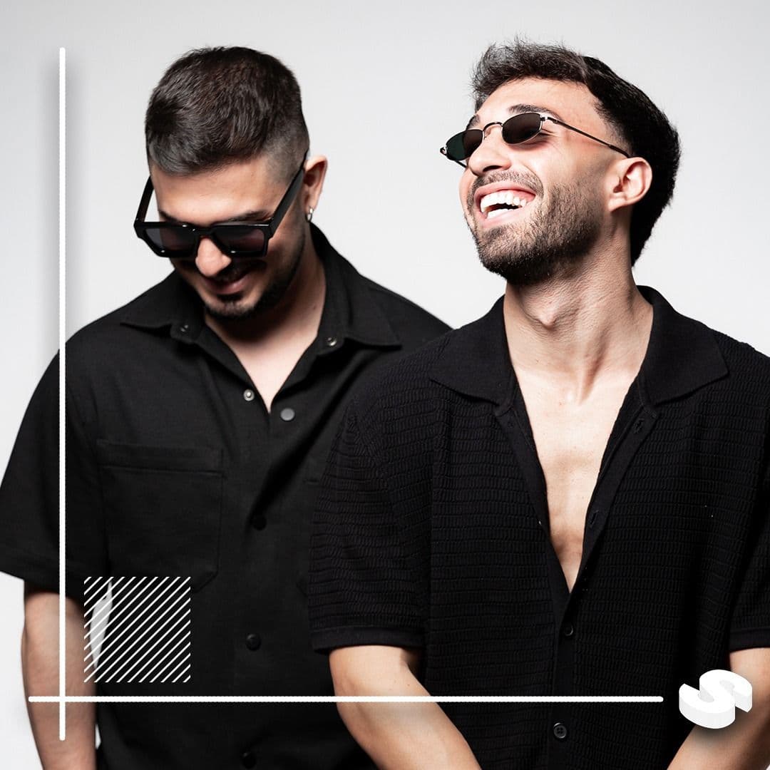 Two men wearing dark shirts and sunglasses, smiling against white background