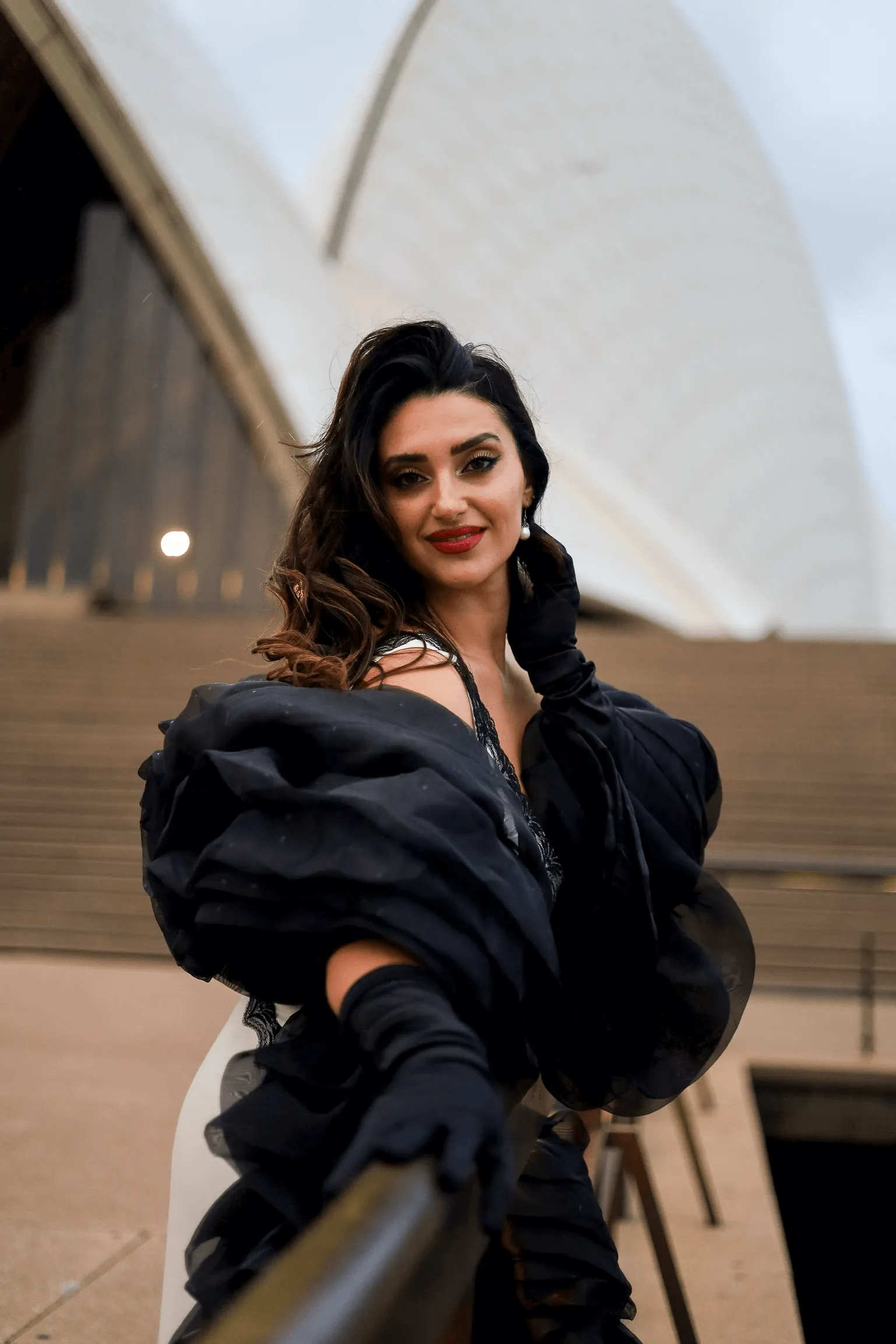 Woman with dark wavy hair and red lipstick wearing black jacket against architectural backdrop.
