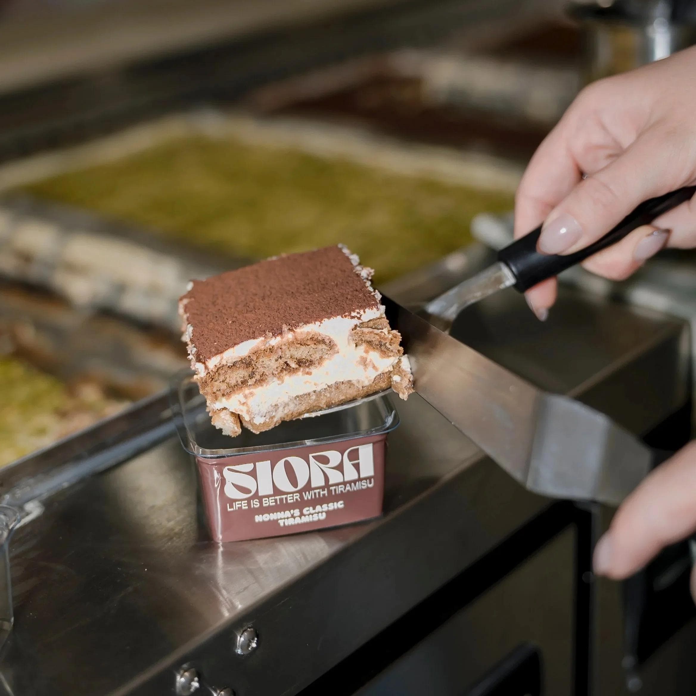 Hand slicing classic tiramisu from Siora branded container at counter