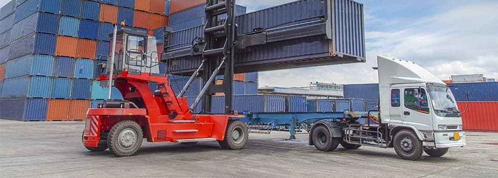 Red forklift lifting shipping container beside truck at container yard