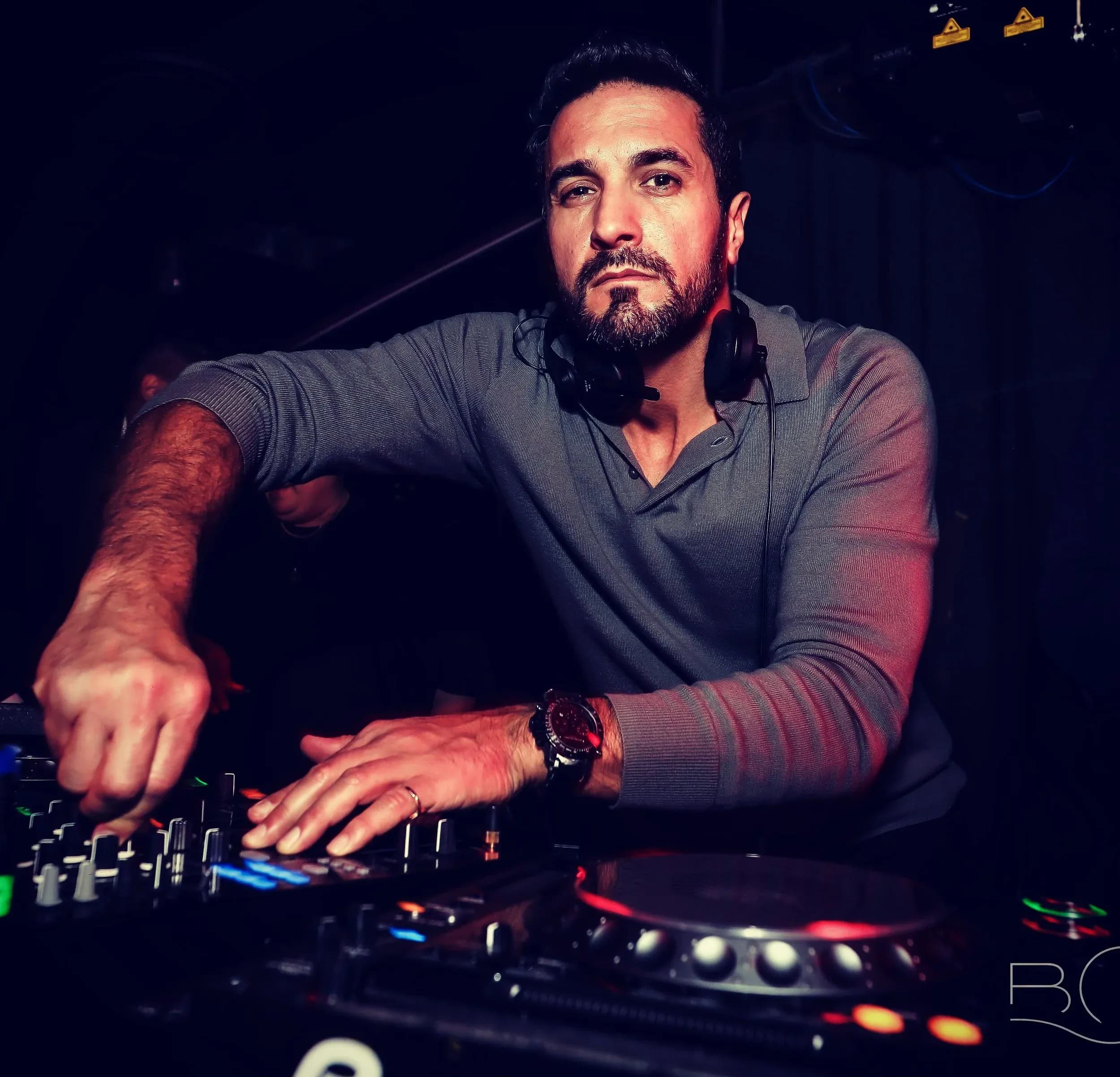 Bearded man wearing headphones at DJ decks, hands on mixer controls, stage lighting.