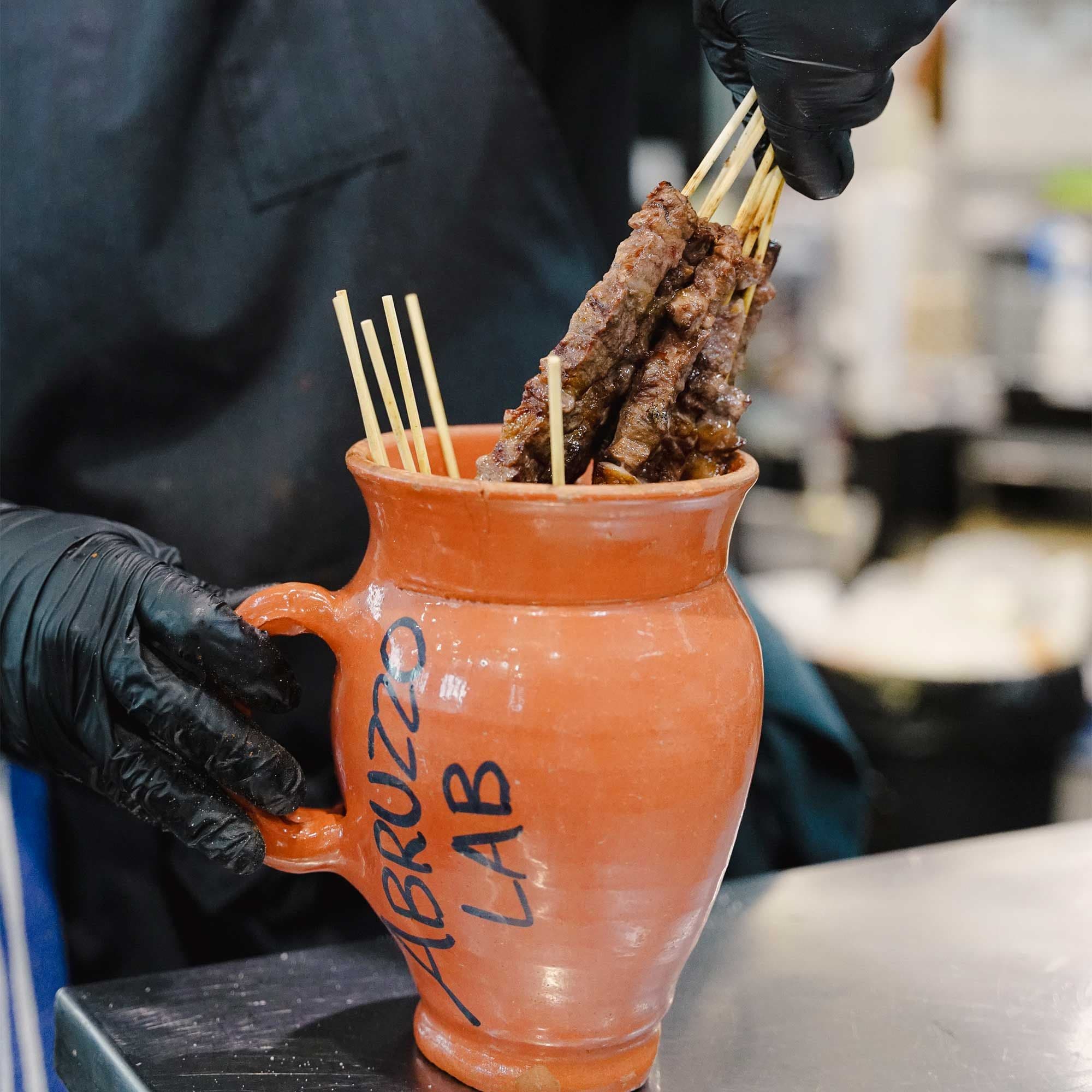 Grilled arrosticini lamb skewers in terracotta jug labeled Abruzzo Lab