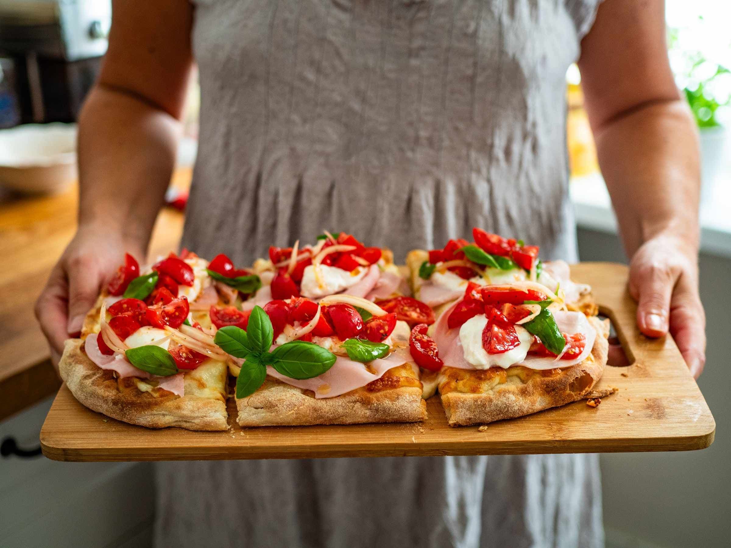 Hands holding wooden board with sliced pinsa topped with ham and tomato