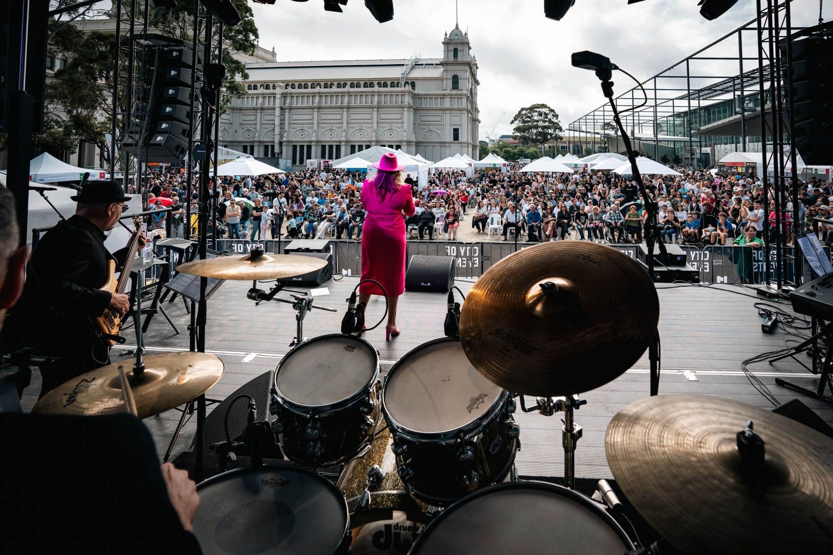 Live jazz performance on main stage with drummer, guitarist, and singer in magenta dress before large crowd