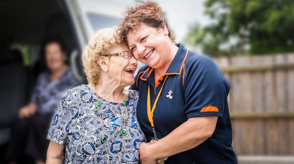 Elderly woman laughing with caregiver in orange-trimmed uniform outdoors
