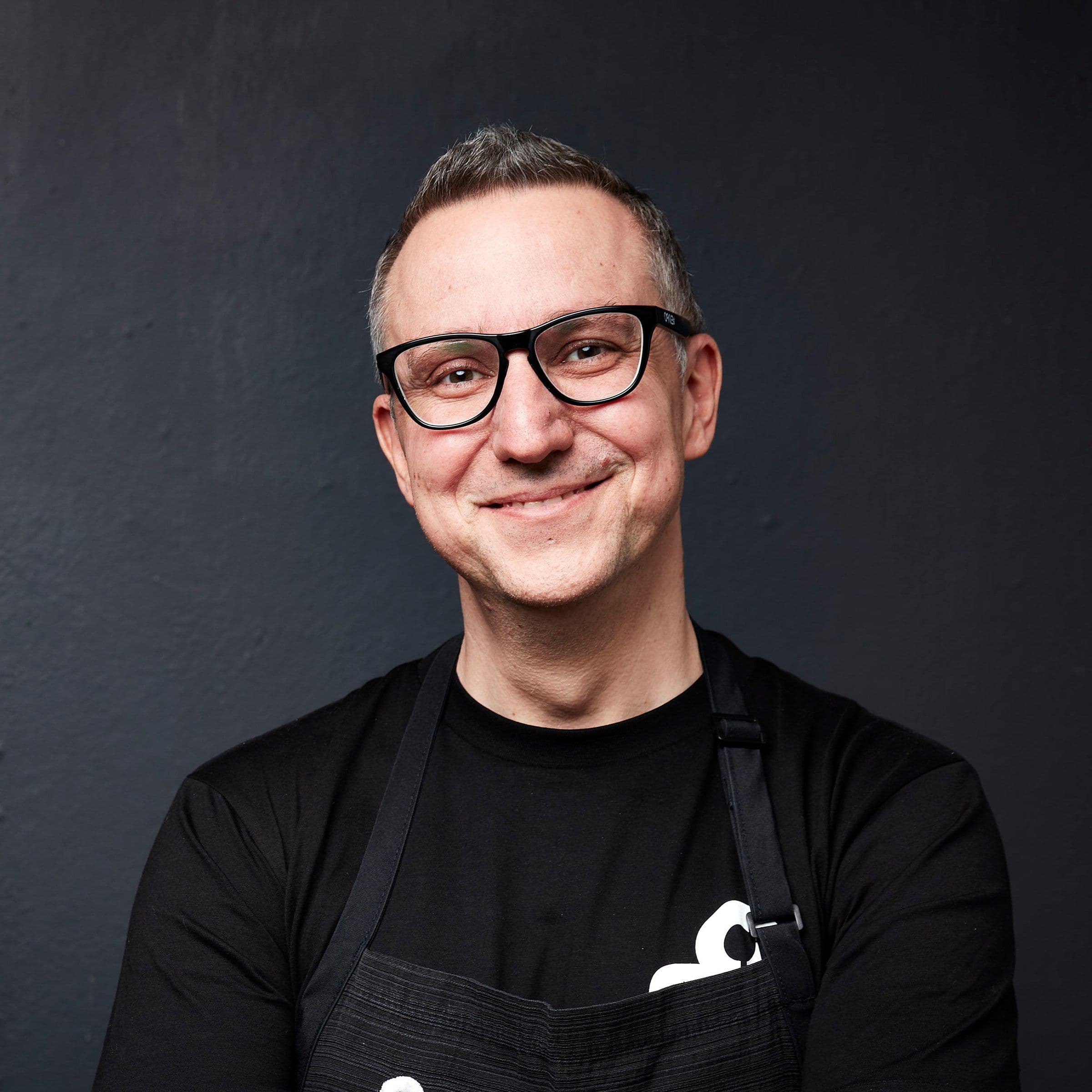 Luca Ciano smiling in glasses and a black apron against a dark background